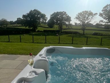 A hot tub with a glass of white wine on the edge overlooking a fenced green field with sheep and trees at Yr Onnen in Bontuchel near Ruthin