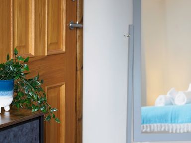 A wooden door next to a table with a green plant in a blue and white pot and a mirror reflecting a bed with a blue blanket and white rolled towels at Trelawney in Mawnan Smith Near Falmouth