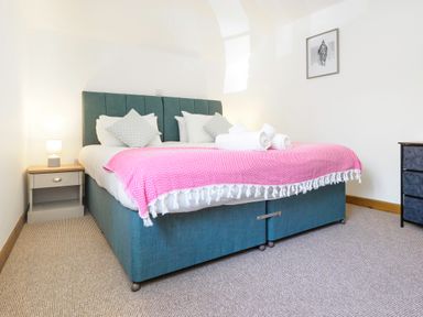 A bedroom with a blue bed, white pillows, a pink throw, two rolled towels, a nightstand with a lamp and a chest of drawers at Trelawney Mawnan Smith Near Falmouth