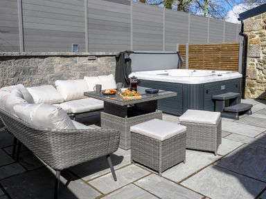 An outdoor patio area with a cushioned wicker sofa set, a table with food and drinks, and a hot tub at Trelawney in Mawnan Smith Near Falmouth