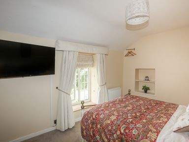 A bedroom with a bed covered with a patterned red bedspread a wall-mounted television a window with white curtains and two shelves with plants at Frongoch Cottage in Red Wharf Bay near Pentraeth