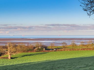 A grassy field with trees and fences overlooking a tidal flat and the sea with ships on the horizon at Frongoch Cottage in Red Wharf Bay near Pentraeth