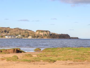 A coastal landscape with sandy beach and green grass facing a bay with houses and a rocky cliff in the distance at Frongoch Cottage in Red Wharf Bay near Pentraeth