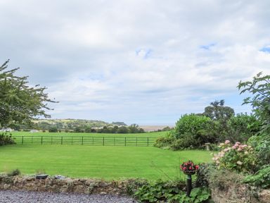 A green lawn with flowerbeds and a stone wall overlooking fields and distant hills at Frongoch Cottage in Red Wharf Bay near Pentraeth