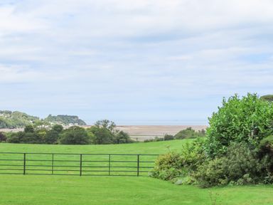 A grassy field with a black metal gate and bushes in the foreground and a sandy beach with trees in the background at Frongoch Cottage Red Wharf Bay near Pentraeth