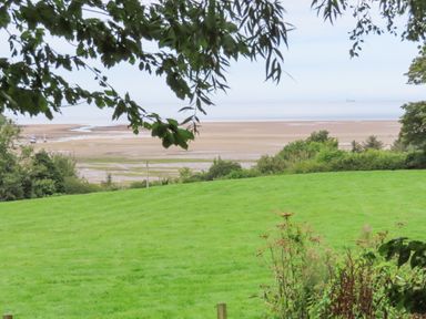 A grassy field with bushes and trees overlooking a sandy beach and sea at Frongoch Cottage in Red Wharf Bay near Pentraeth