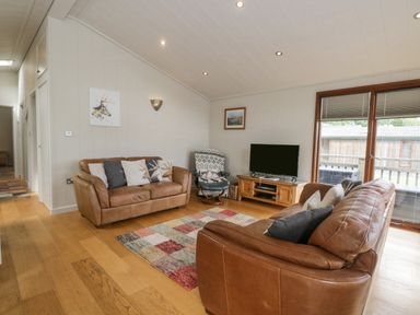 A living room with two brown leather sofas, a patterned rug, a TV on a wooden stand, and a window with blinds at Rivendell Lodge in Roger Ground near Hawkshead