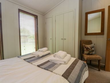 A bedroom with a bed, folded towels on a striped blanket, a wicker chair with a pillow, a wall mirror, and a window with wooden blinds at Rivendell Lodge Roger Ground near Hawkshead