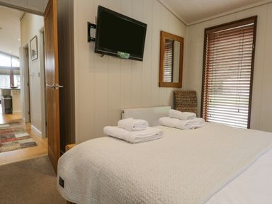A bedroom with a bed folded towels a wall-mounted tv a mirror a chair and window blinds at Rivendell Lodge Roger Ground near Hawkshead