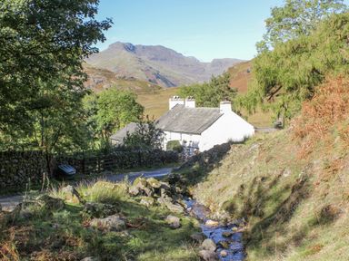 A white house near a stream with trees and mountains in the background at Rivendell Lodge in Roger Ground near Hawkshead