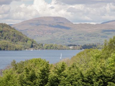 A lake with a sailboat in the middle surrounded by green trees and hills under a partly cloudy sky at Rivendell Lodge in Roger Ground near Hawkshead