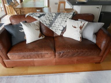 A brown leather sofa with four cushions and a patterned throw in a living area with a wooden dining table and chairs at Rivendell Lodge Roger Ground near Hawkshead