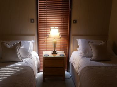 A bedroom with two single beds separated by a wooden nightstand with a lamp in front of a window with closed blinds at Rivendell Lodge Roger Ground near Hawkshead