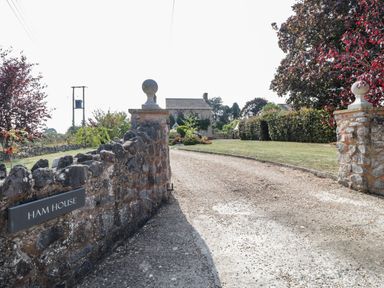 A stone wall and gateposts leading to a gravel driveway with a house and garden in the background at The Forge in Taunton
