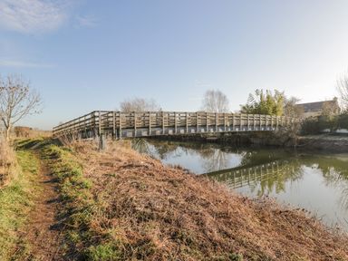 A wooden bridge over a river with a grassy path alongside and trees in the background at The Forge in Taunton