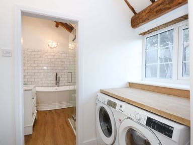 A utility room with a washing machine and dryer under a wooden countertop next to a window and a bathroom with a bathtub visible through an open door at Well Cottage in Grimston