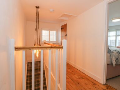 A hallway with wooden floor and white railing leading to stairs with plaid carpet and a bedroom visible through a doorway at Well Cottage in Grimston