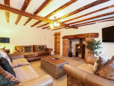 A living room with wooden beams on the ceiling two brown sofas a wooden coffee table a fireplace with a wood-burning stove and a wall-mounted TV at Well Cottage in Grimston