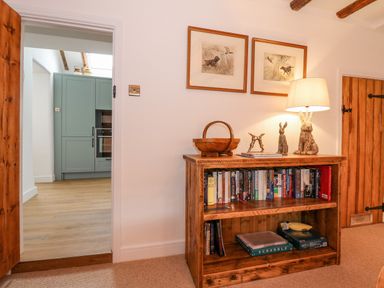 A wooden bookshelf with books and board games under a lamp with figurines and framed pictures on the wall in a room at Well Cottage in Grimston