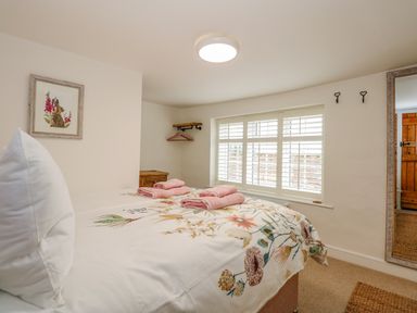 A bedroom with a floral bedspread and pink towels on the bed a side table under a corner shelf with hangers a large window with white shutters a full length mirror and a framed rabbit picture on the wall at Well Cottage in Grimston