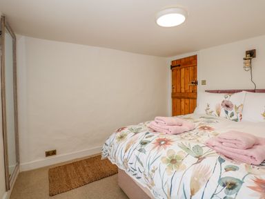 A bedroom with a floral patterned bedspread on a bed two folded pink towels on the bed a wooden door a woven rug and a wall-mounted light at Well Cottage in Grimston