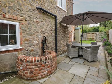 An outdoor patio area with stone wall a brick water pump a round table with an umbrella and chairs at Well Cottage in Grimston