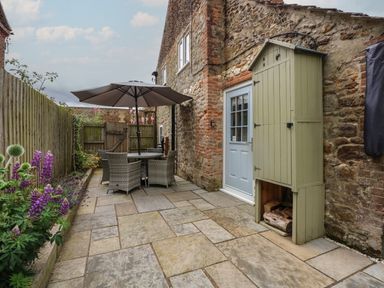 A stone patio with outdoor dining furniture and umbrella next to a stone wall with a blue door and green storage unit with logs in Grimston