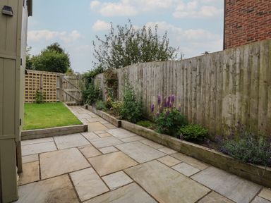A paved patio with flower beds and a grassy area enclosed by wooden fencing at Well Cottage in Grimston