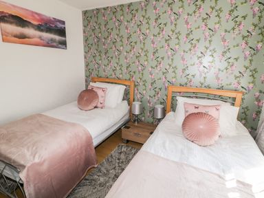 A bedroom with two single beds with pink cushions and blankets a bedside table with lamps and floral wallpaper at Peters Farmhouse in Cookstown