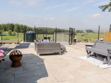 An outdoor patio with grey chairs and table a grey bench and a fire pit with a fenced garden and trees in the background at Peters Farmhouse in Cookstown