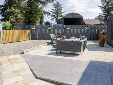 An outdoor patio with grey and beige tiles a grey wicker sofa set a wooden chair potted flowers a wooden fence and trees at Peters Farmhouse in Cookstown
