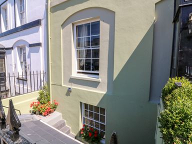 An exterior view of a light green building with two white-framed windows and flowers in pots near the windows at Drake's Retreat in Plymouth