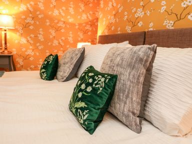 A bed with white sheets and decorative green and brown pillows with floral wallpaper and a lamp in the background at Drakes Retreat in Plymouth