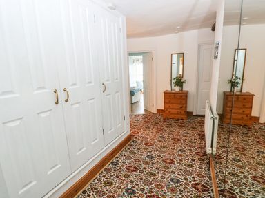 A hallway with patterned carpet a wooden chest of drawers with mirror and plants and white built in cupboards and a large wall mirror at Gower View in Tenby