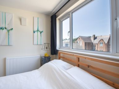 A bedroom with a wooden bed frame white bedding two abstract paintings a radiator and a window with a view of brick houses at Ayla in Lyme Regis