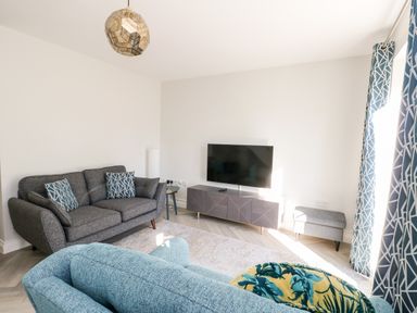 A living room with a grey sofa, blue armchair, TV on a stand, and blue patterned curtains at 3 Trem Y Dolydd Dolgarrog near Llanrwst