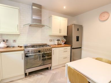 A kitchen with white cabinets a stainless steel stove and refrigerator a white table with wooden chairs and a pink clock on the wall at 3 Trem Y Dolydd in Dolgarrog near Llanrwst