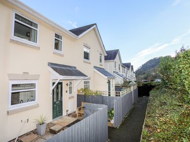 A row of beige two story houses with slate roofs and green doors along a narrow paved path with wooden fences and shrubbery at 3 Trem Y Dolydd in Dolgarrog near Llanrwst