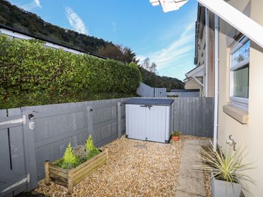 An outdoor area with gravel ground a wooden planter with plants a gray fence a gray storage box and a beige building with a window at 3 Trem Y Dolydd Dolgarrog near Llanrwst
