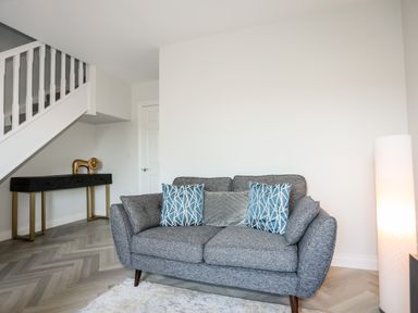 A grey sofa with patterned and plain cushions next to a lit floor lamp and a console table under white stairs at 3 Trem Y Dolydd in Dolgarrog near Llanrwst