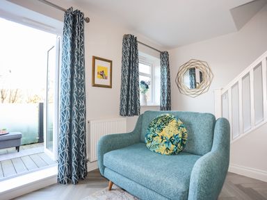 A small teal sofa with a round floral cushion next to a window and glass door with blue patterned curtains at 3 Trem Y Dolydd in Dolgarrog near Llanrwst