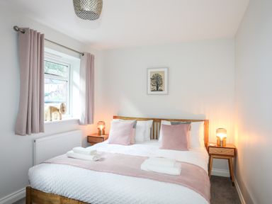 A bedroom with a wooden bed pink pillows bedside tables with lamps a window with curtains and a framed picture on the wall at 3 Trem Y Dolydd in Dolgarrog near Llanrwst