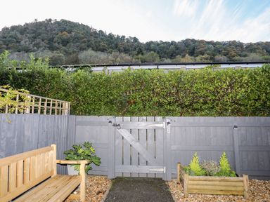 An outdoor patio area with a wooden bench, a gray wooden fence with a gate, green plants, and a hill with trees in the background at 3 Trem Y Dolydd in Dolgarrog near Llanrwst