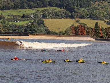 People surfing and lying on surfboards in a man made wave pool with hills and trees in the background at 3 Trem Y Dolydd in Dolgarrog near Llanrwst