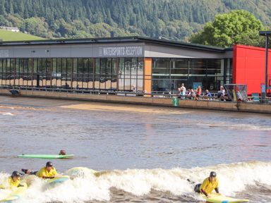 A watersports reception building with people sitting outside and three people with surfboards in the water at 3 Trem Y Dolydd in Dolgarrog near Llanrwst