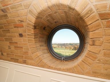 A circular window set in a thick brick wall with a view of green fields and trees at Tan y Wawr- Onnen in Llanfair Caereinion