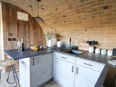 A kitchen with brick walls a black countertop white cabinets a sink a fruit basket and bar stools at Tan y Wawr- Onnen in Llanfair Caereinion