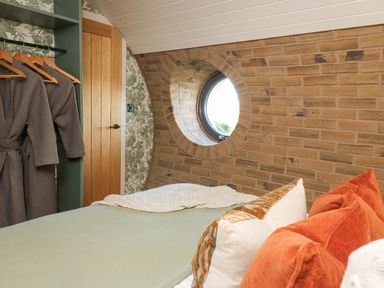 A bedroom with a green bedspread orange pillows a round window brick wall wooden door and robes hanging in an open closet at Tan y Wawr- Onnen in Llanfair Caereinion