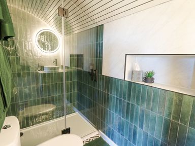 A bathroom with green tiled walls a glass shower door a round illuminated mirror and a shelf with a plant and tissue box at Tan y Wawr- Onnen in Llanfair Caereinion