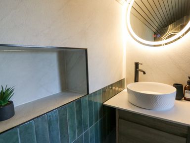 A bathroom with a round mirror with lights a white sink with black faucet green tiles and a small plant at Tan y Wawr- Onnen in Llanfair Caereinion